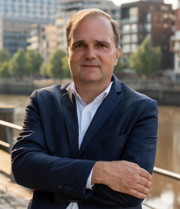 Portrait Patrick von der Born Mann in Anzug mit verschränkten Armen, Hintergrund mit Wasser und Gebäuden.
